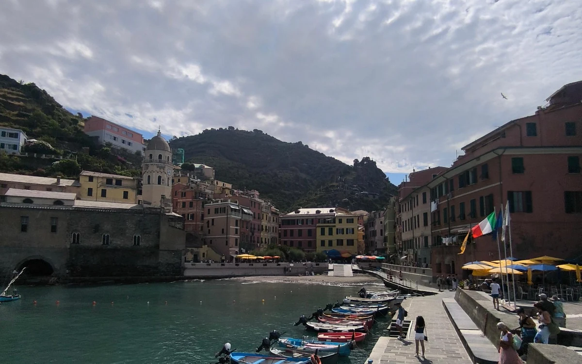 Small boats in a harbour below pastel houses and a church tower, green hills behind