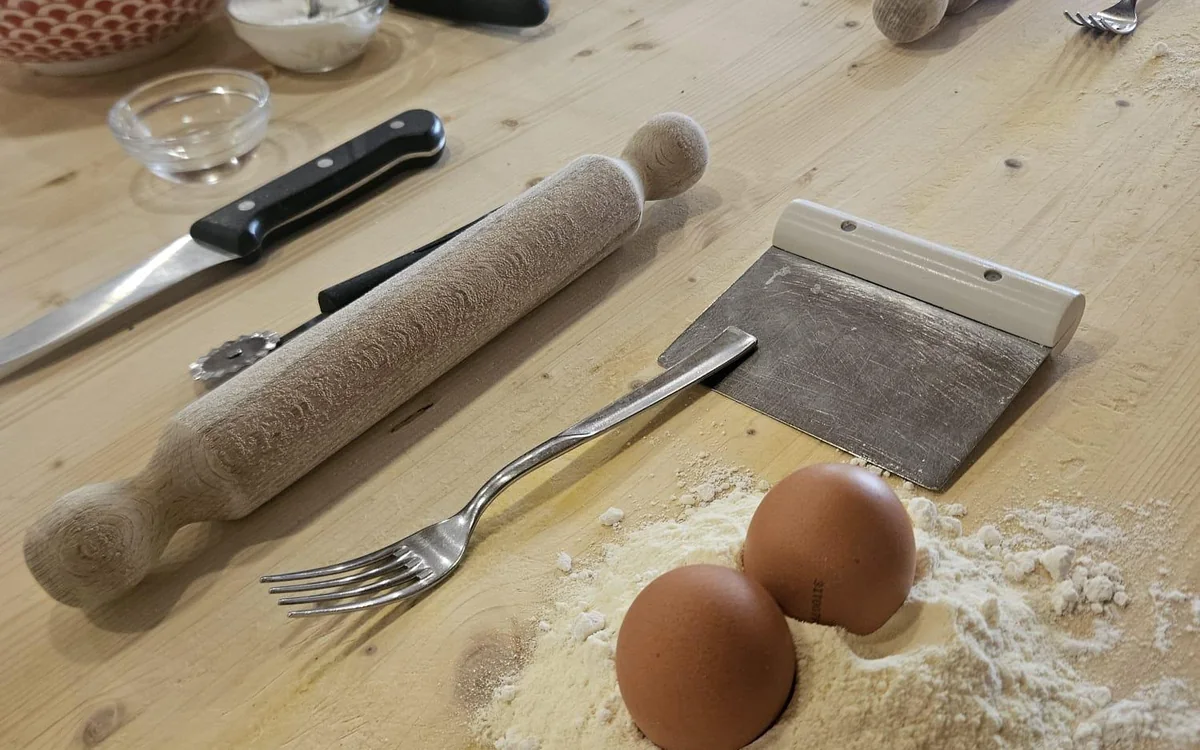 Two eggs in a mound of flour beside a wooden rolling pin, a fork and a dough scraper