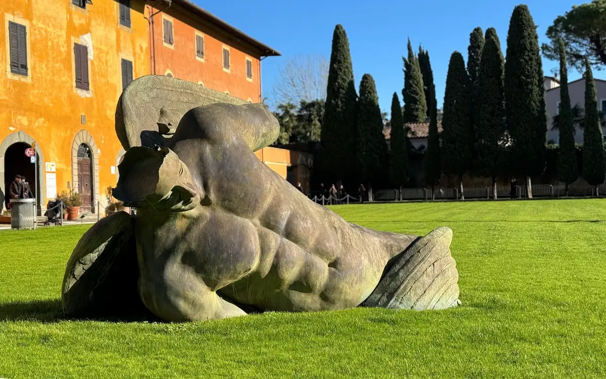 A large bronze figure with wings lying on the lawn in Pisa, an orange house and cypresses behind