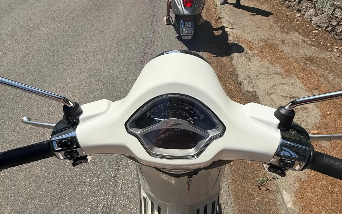 Looking down over the handlebars and speedometer of a white Vespa, another Vespa on the road ahead