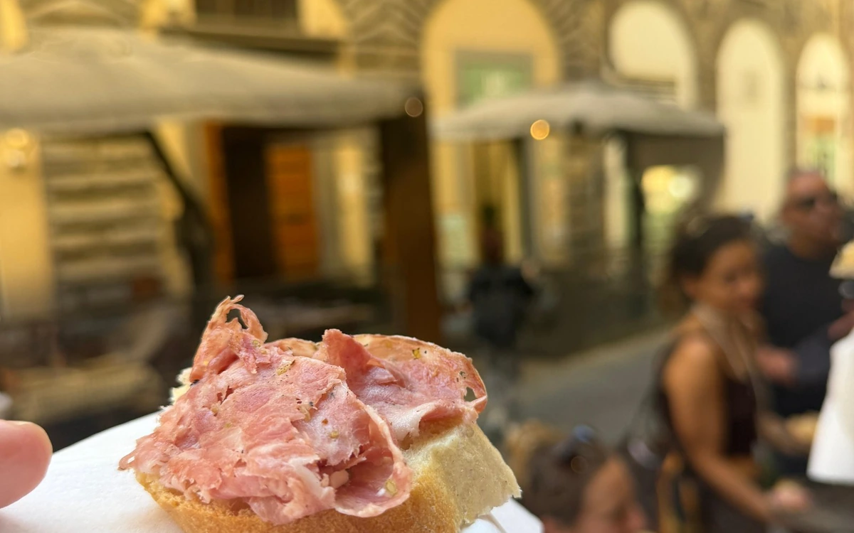 Thin slices of cured meat on a piece of bread, held up in a Florence street with arcades and people passing behind