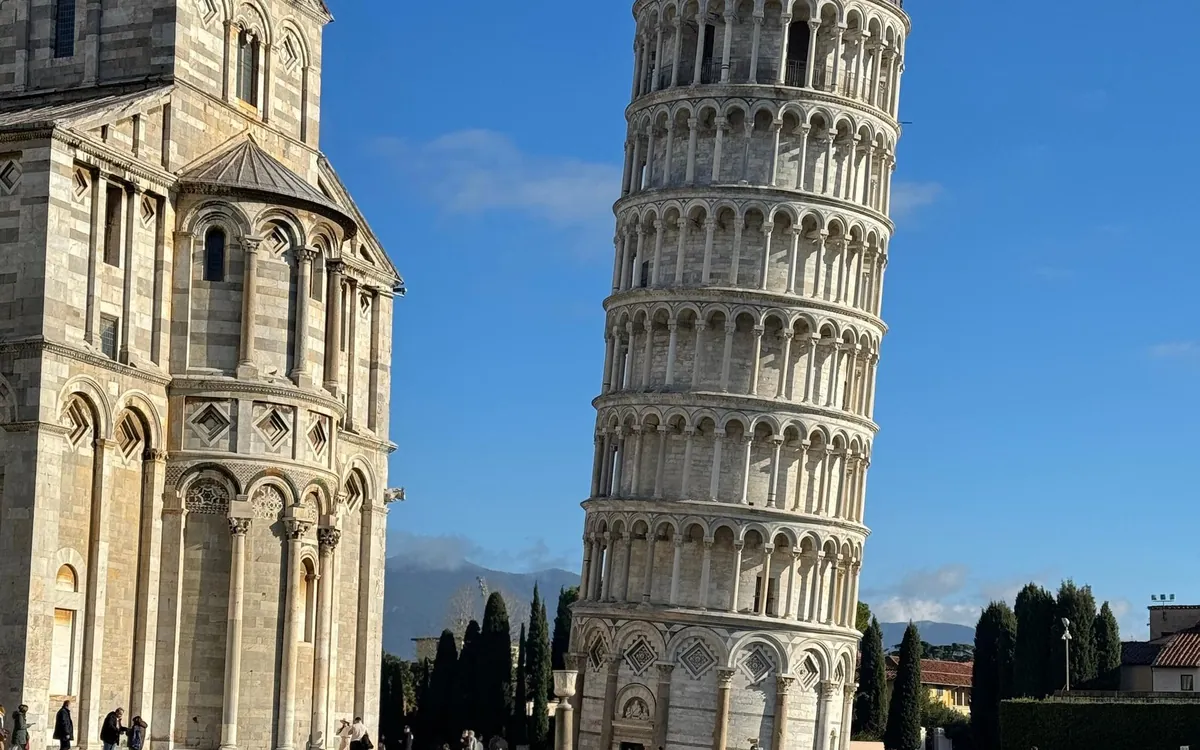 The Leaning Tower of Pisa beside the apse of the cathedral under a blue sky, cypresses below